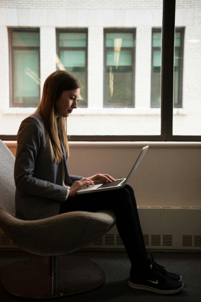 Woman sitting on a chair using a laptop by the window, engaged in remote work.