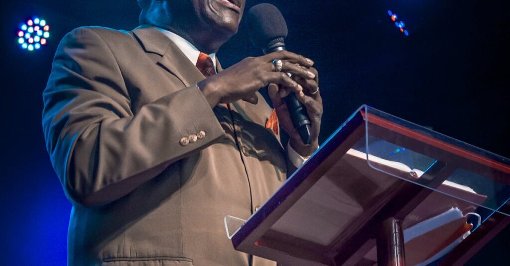 An African American man passionately speaks into a microphone while standing at a podium under bright stage lights.